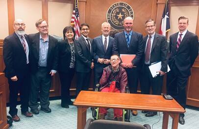 Group of people in suits and a wheelchair user in an office standing and sitting in front of the state seal of Texas and the US and Texas flags