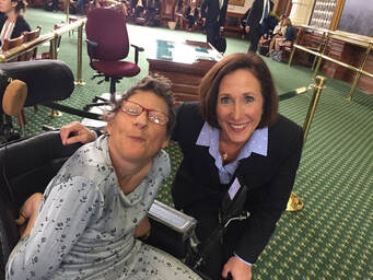 Image of white woman wearing red glasses and using a wheelchair alongside a white woman in a suit
